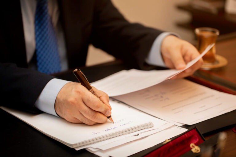 Uomo in giacca e cravatta scrive su un blocco note, unendo documenti.