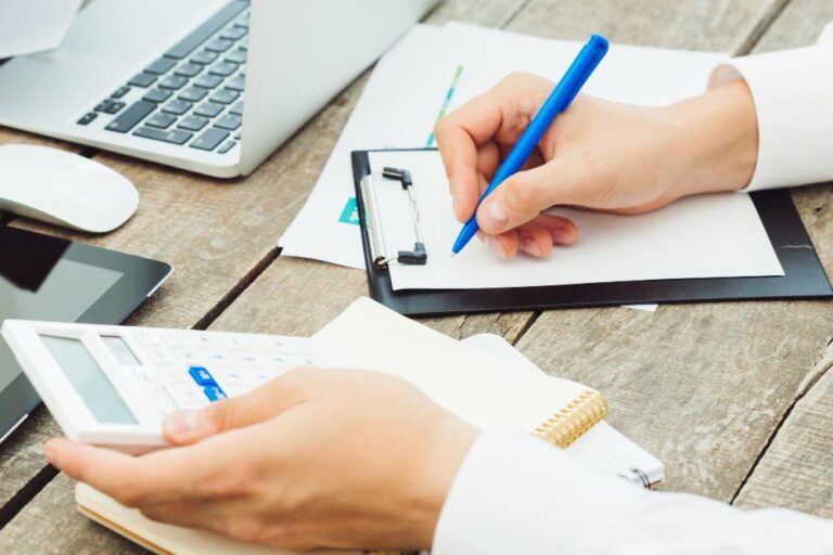 Mani su una scrivania in legno lavorano con laptop, calcolatrice e blocco note.