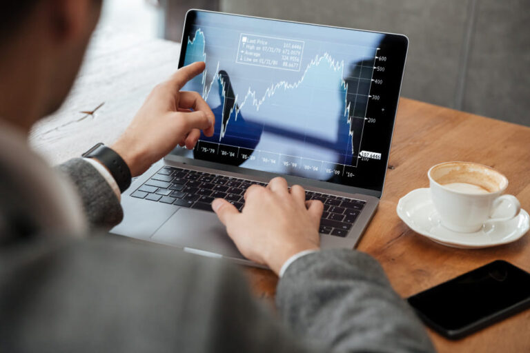 Uomo in giacca grigia lavora al laptop, puntando grafico a linea, con caffè e telefono.